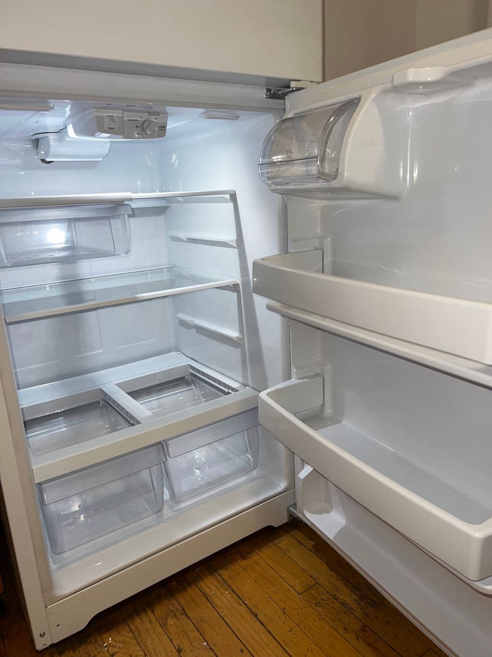 Empty refrigerator interior with transparent shelves and drawers on wooden floor.