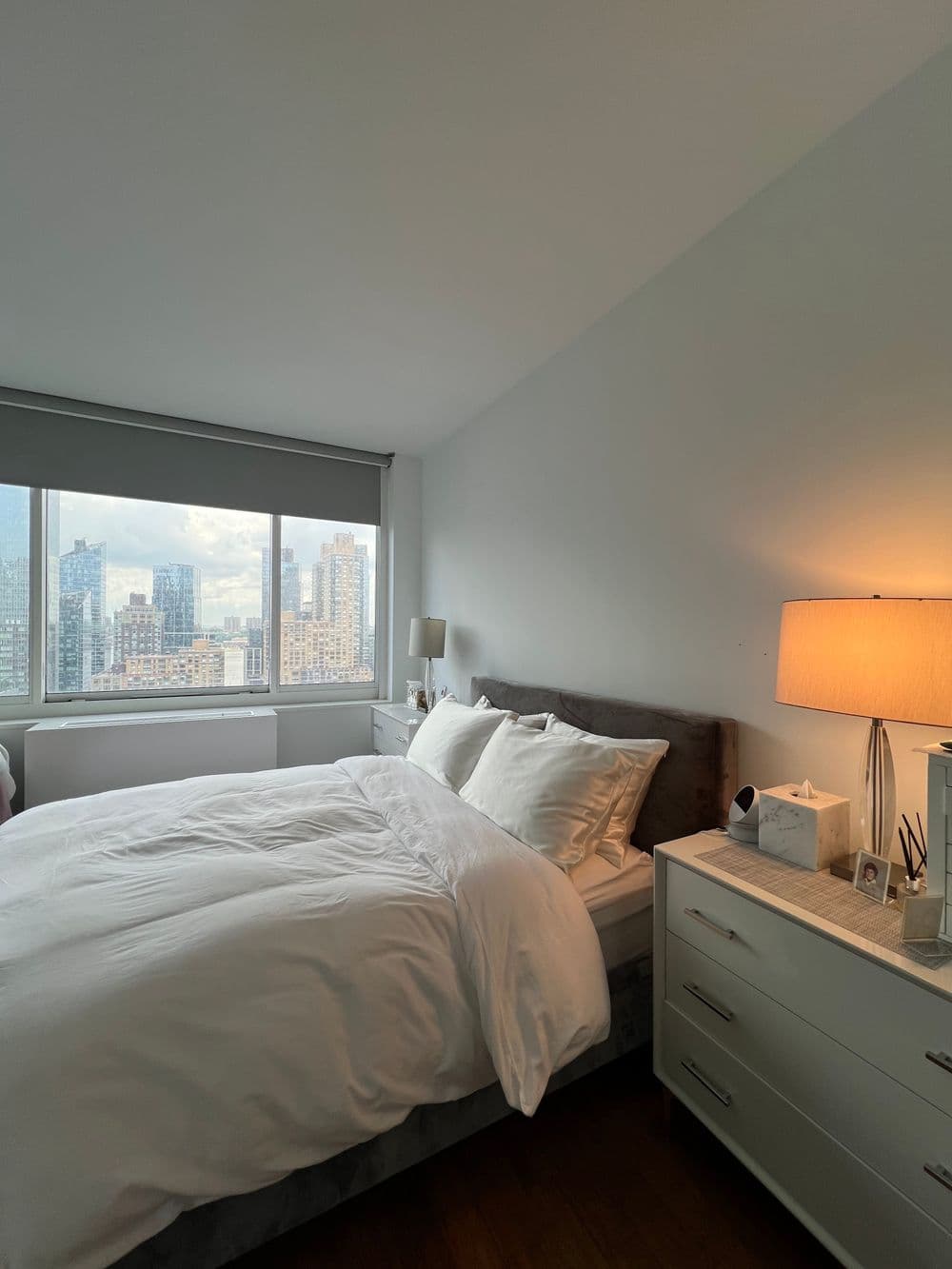 Cozy bedroom with gray upholstered bed, modern nightstand, and city skyline view.