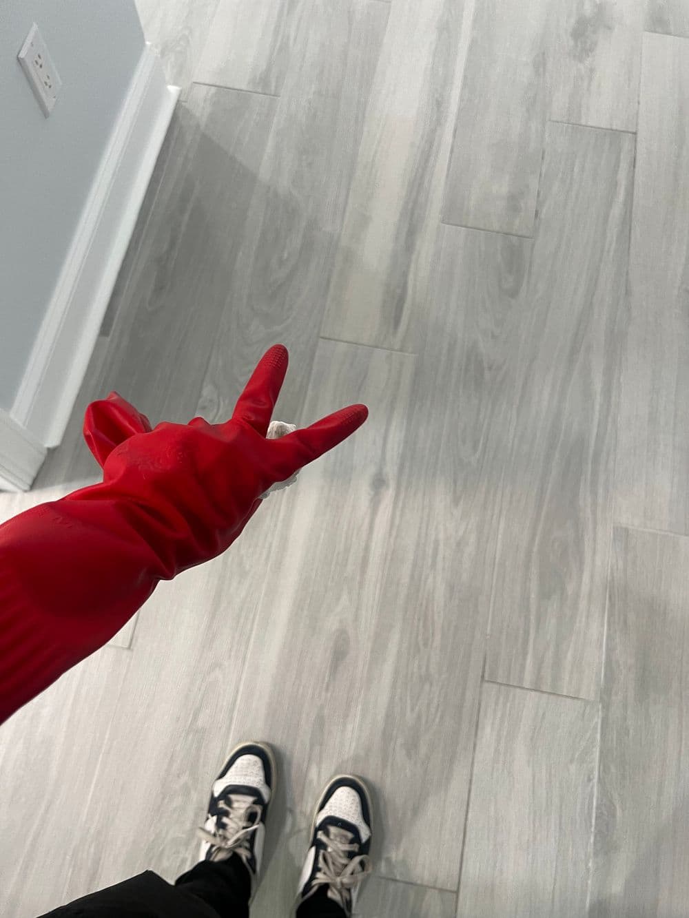 Red rubber glove making a peace sign over a light wooden floor with sneakers visible.