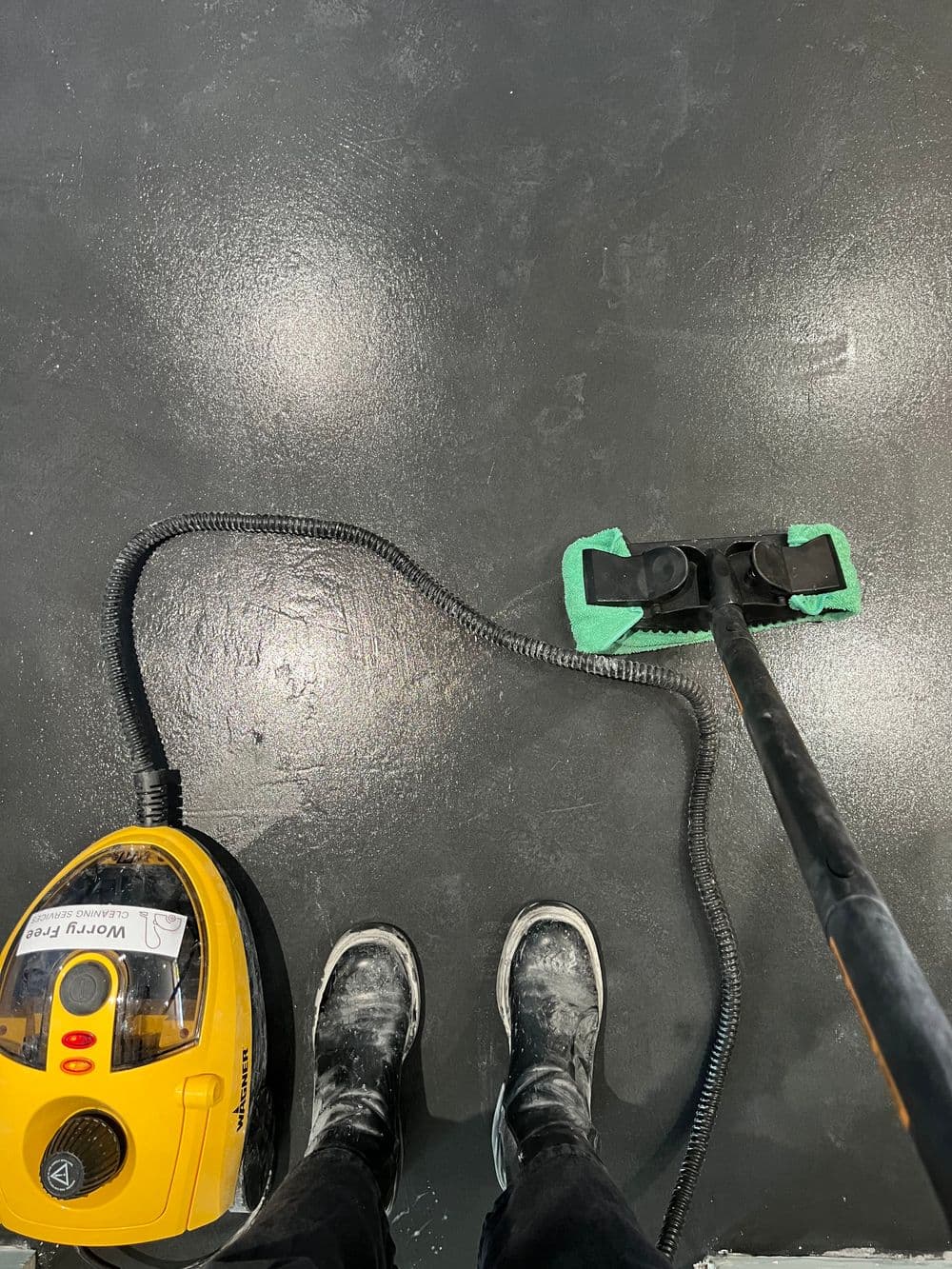 Person cleaning a dark floor with a steam cleaner and wearing dusty shoes.